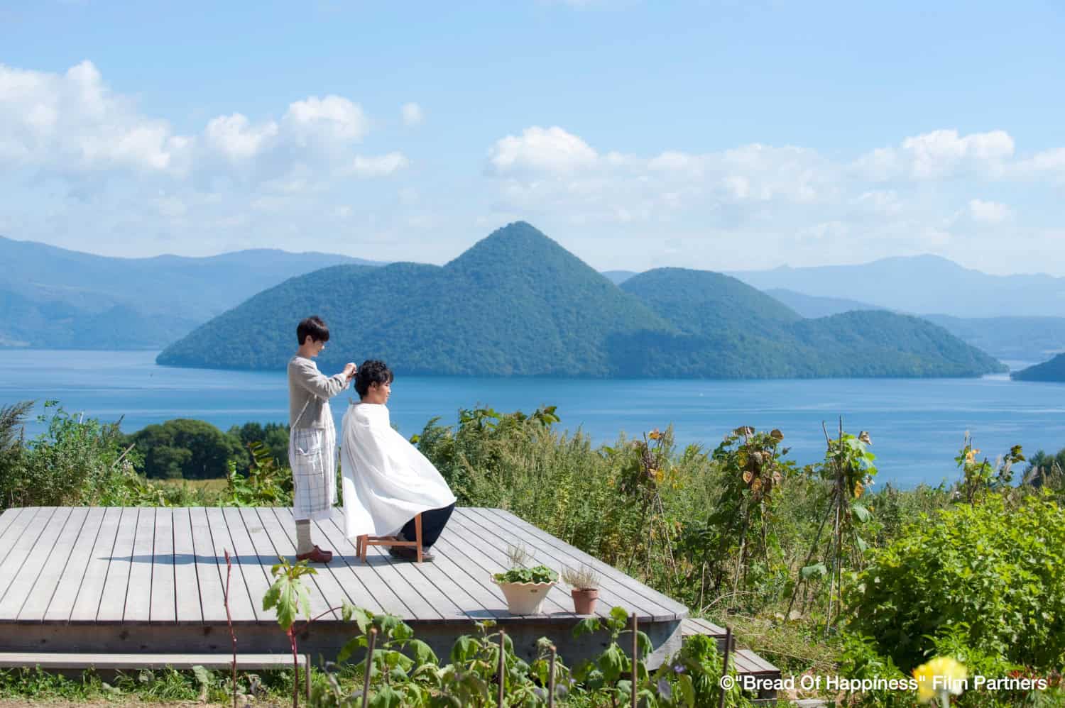 Movie still for Bread of Happiness showing a sea view overlooking mountains and two people, one cutting the other's hair