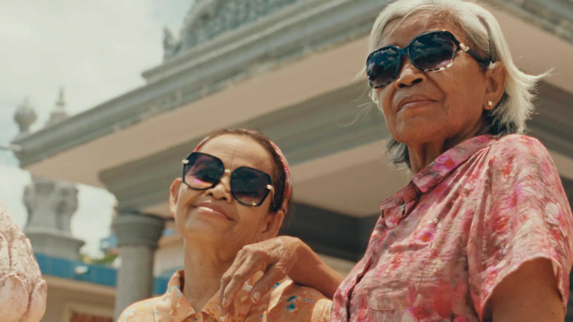 Two fashionable grandmothers lean against each other in a sundrenched gravesite.